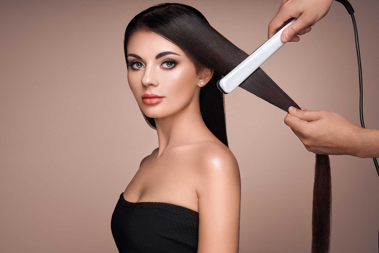 A woman with long, smooth brown hair is getting her hair straightened with a flat iron. She wears a black top, exuding elegance and poise.